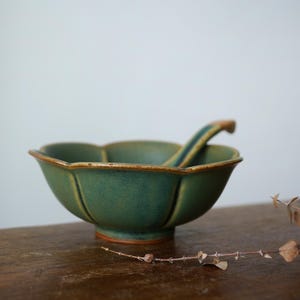 Może przedstawiać: Zielona ceramiczna miska z pasującą łyżką. Miska ma unikalną, karbowaną krawędź i teksturowaną powierzchnię. Łyżka spoczywa wewnątrz miski. Miska i łyżka są w kolorze ciemnozielonym z brązową obw&oacute;dką i podstawą.