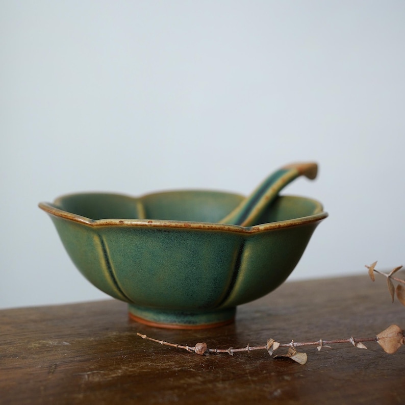 Może przedstawiać: Zielona ceramiczna miska z pasującą łyżką. Miska ma unikalną, karbowaną krawędź i teksturowaną powierzchnię. Łyżka spoczywa wewnątrz miski. Miska i łyżka są w kolorze ciemnozielonym z brązową obw&oacute;dką i podstawą.