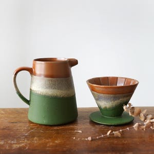 May include: A ceramic coffee pour-over set with a matching pitcher. The pitcher is brown at the top, transitioning to a light color, then green. The pour-over has a similar color scheme. The set is on a wooden surface.