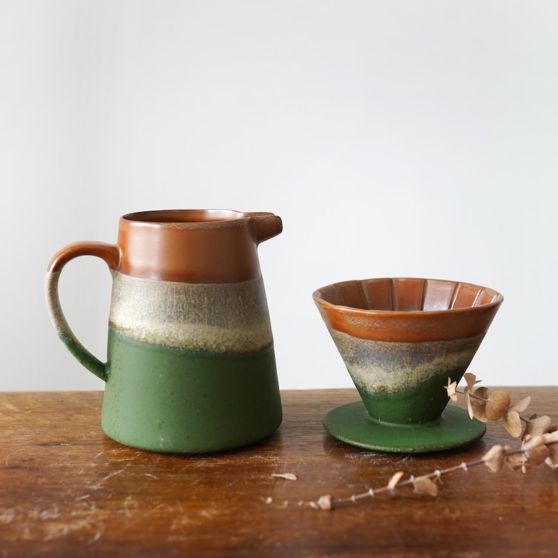 May include: A ceramic coffee pour-over set with a matching pitcher. The pitcher is brown at the top, transitioning to a light color, then green. The pour-over has a similar color scheme. The set is on a wooden surface.