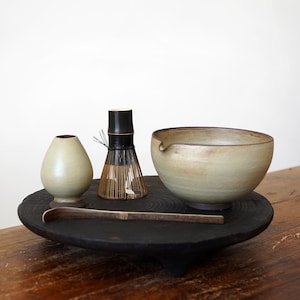 Puede incluir: Un juego de té matcha sobre una bandeja de madera oscura. El juego incluye un cuenco de cerámica, un batidor de bambú, un pequeño recipiente de cerámica y una cuchara de bambú. El cuenco es de color beige claro y los demás artículos son de tonos de madera natural.
