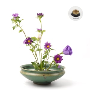 Könnte beinhalten: Eine grüne Keramik-Blumenschale mit wassergefülltem Innenraum, in der lila und gelbe Blumen stehen. Eine schwarze und goldene Blumenfrosch-Halterung ist enthalten. Die Schale hat ein breites, flaches Design mit einem dezenten, strukturierten Finish.