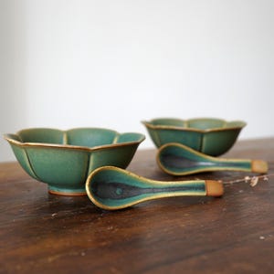 Może przedstawiać: Zestaw ręcznie robionych ceramicznych misek i łyżek. Miski i łyżki są w kolorze głębokiego, morskozielonego z złotym obramowaniem. Łyżki mają czarne środki i drewniane uchwyty. Miski mają unikalny kształt płatka.