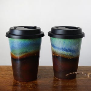 May include: Two ceramic to-go mugs with black lids. The mugs have a blue, green, and brown glaze with a layered effect.