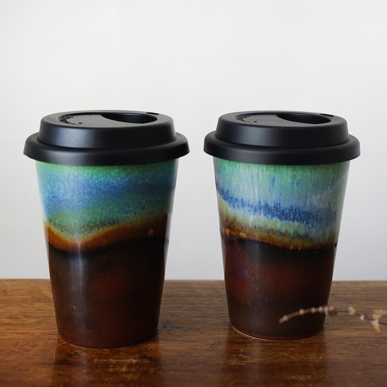 May include: Two ceramic to-go mugs with black lids. The mugs have a blue, green, and brown glaze with a layered effect.