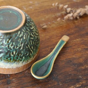 May include: A ceramic bowl and a matching spoon featuring a textured, mottled glaze in shades of deep forest green, dark charcoal, and earthy tan. The bowl has a rounded shape with a visible base, while the spoon has a long, slender handle.