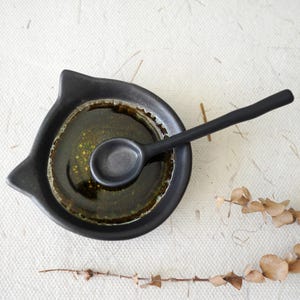 May include: A black ceramic sauce dish with a small spoon. The dish contains a dark liquid with visible specks. The dish has a unique shape with two pointed edges. The spoon rests inside the dish.