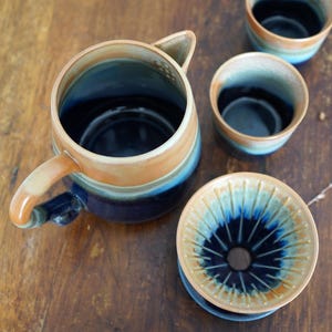 May include: A ceramic coffee set with a tan, blue, and brown glaze. The set includes a coffee server with a handle and spout, a coffee dripper, and two small cups. The pieces are arranged on a wooden surface.
