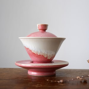 May include: A ceramic gaiwan tea set with a lid, bowl, and saucer. The bowl is a light pink with a white and pink gradient. The set is on a wooden surface.