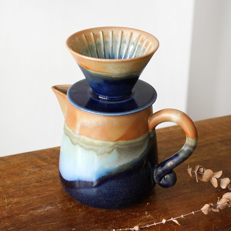 May include: A ceramic pour-over coffee maker with a matching dripper. The coffee maker is a mix of blue, orange, and cream colors. The dripper is a light brown color with vertical grooves. The handle is a curved design.