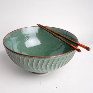 May include: A green ceramic bowl with a textured, ribbed exterior. The bowl is empty and has a pair of wooden chopsticks resting on the rim.