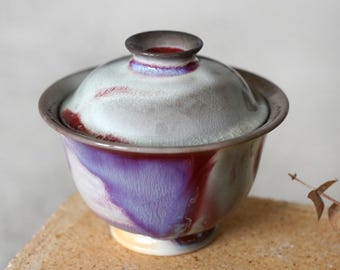 Puede incluir: Un cuenco de té gaiwan de cerámica con tapa a juego, que presenta un esmalte reactivo en tonos púrpura intenso, rojo carmesí y gris suave. El cuenco tiene un borde ancho y acampanado, un cuerpo redondeado y una base pequeña con acabado brillante.