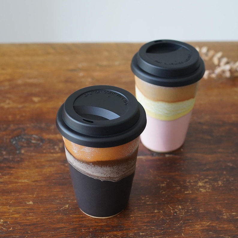 May include: Two ceramic travel mugs with black lids. One mug has a color gradient of black, brown, and tan. The other mug has a color gradient of pink, yellow, and brown. Both mugs are on a wooden surface.