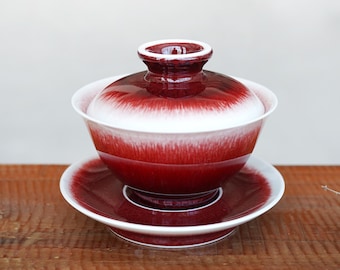 Puede incluir: Un juego de té gaiwan de cerámica que consta de un cuenco, una tapa y un platillo. El conjunto presenta un esmalte rojo intenso que se transforma en una banda blanca brillante alrededor del centro del cuenco y la parte superior de la tapa.