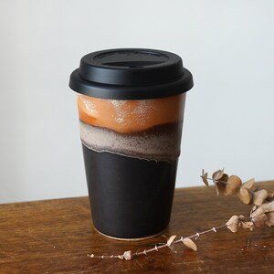 May include: A black travel mug with a black lid. The mug has a unique design with layers of color, including orange, brown, and gray. The mug is sitting on a wooden surface.