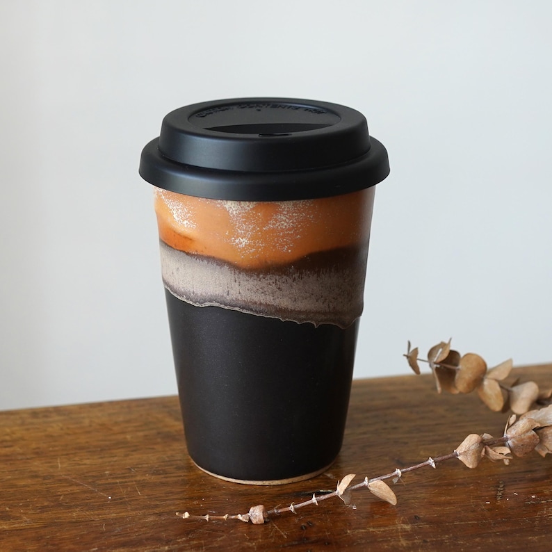 May include: A black travel mug with a black lid. The mug has a unique design with layers of color, including orange, brown, and gray. The mug is sitting on a wooden surface.