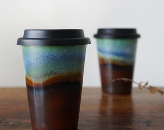 May include: Two ceramic travel mugs with black lids. The mugs have a blue, green, and brown glaze with a layered effect. The mugs are sitting on a wooden surface.