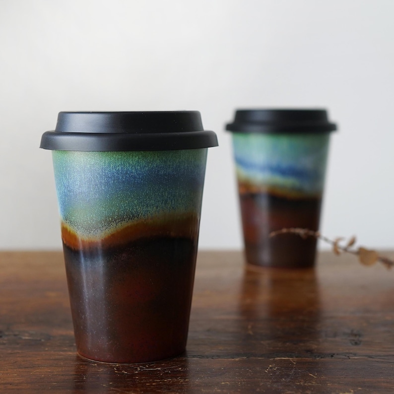 May include: Two ceramic travel mugs with black lids. The mugs have a blue, green, and brown glaze with a layered effect. The mugs are sitting on a wooden surface.