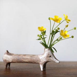 Pode incluir: Um vaso de cerâmica em forma de dachshund com um esmalte marrom e branco, contendo um buquê de flores amarelas.