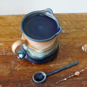 May include: A handmade ceramic pitcher with a dark blue interior and a spout. The exterior features bands of color in shades of blue, orange, and green. A matching blue ceramic spoon rests beside the pitcher on a wooden surface.