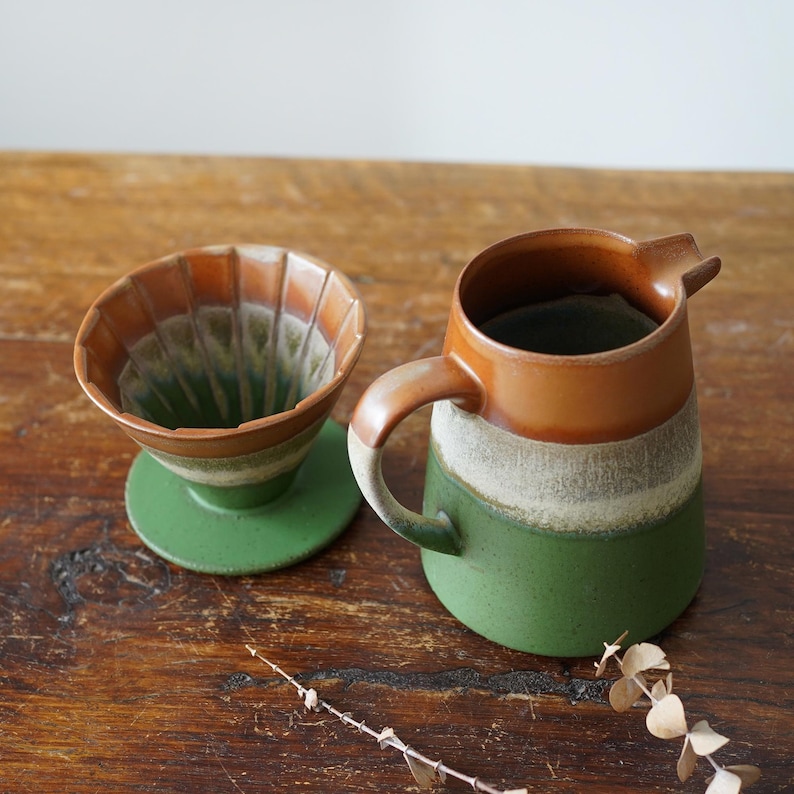 May include: A ceramic coffee pour-over set and pitcher. The pour-over has a brown and green design, with a matching green base. The pitcher is brown, white, and green with a handle and spout. The set is on a wooden surface.