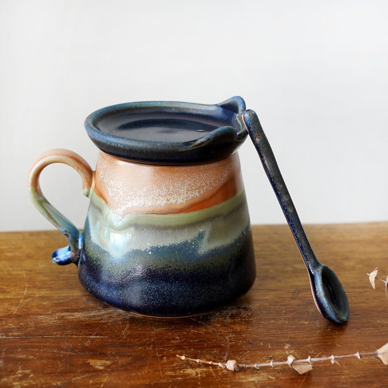 May include: A handmade ceramic mug with a lid and spoon. The mug features a color gradient from tan to blue, with a curved handle. The spoon and lid are dark blue. The mug is sitting on a wooden surface.