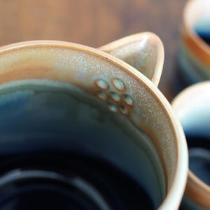 May include: Close-up of a ceramic mug with a unique design. The mug features a gradient of colors, including blue, brown, and cream. The rim has a textured, speckled appearance. The handle has a series of small, circular indentations. The mug is part of a set.