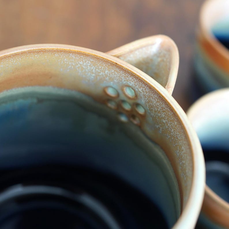May include: Close-up of a ceramic mug with a unique design. The mug features a gradient of colors, including blue, brown, and cream. The rim has a textured, speckled appearance. The handle has a series of small, circular indentations. The mug is part of a set.