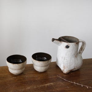 Op de afbeelding: Een keramische theeset met een wit en bruin gespikkelde afwerking. De set bevat een kan met een tuit en handvat, en twee kleine kopjes. De binnenkant van de kopjes is donkerbruin.