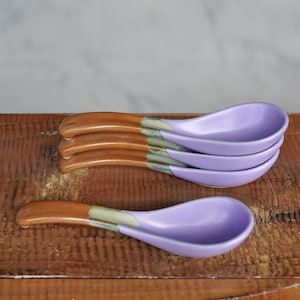 May include: A set of four ceramic soup spoons featuring a smooth, lavender-colored bowl and a contrasting brown handle. The transition between the lavender bowl and the brown handle is marked by a subtle, muted green band of glaze on each spoon.