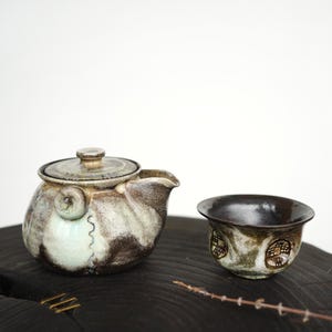 May include: A ceramic teapot with a lid and a matching small cup, both featuring a mottled glaze in shades of cream, brown, and dark grey. The teapot has a rounded body with a side handle and a spout, while the cup has a dark interior.