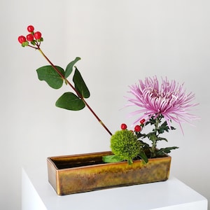 Può includere: Composizione floreale con un crisantemo viola chiaro, bacche verdi e un fiore verde spinoso. La composizione è in un vaso rettangolare in ceramica color bronzo. Lo sfondo è bianco.