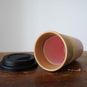 May include: A ceramic travel mug with a pink interior and a black lid. The mug has a light brown exterior with a yellow stripe. The mug is lying on its side on a wooden surface.
