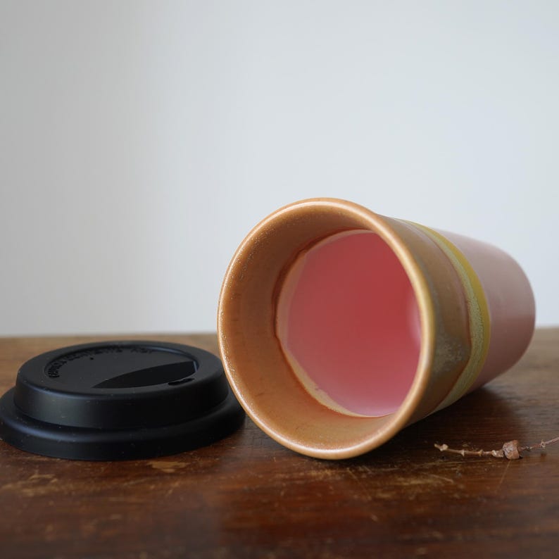 May include: A ceramic travel mug with a pink interior and a black lid. The mug has a light brown exterior with a yellow stripe. The mug is lying on its side on a wooden surface.