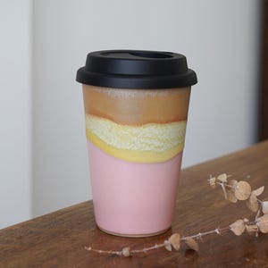 May include: A ceramic travel mug with a black lid. The mug has a pink base, a yellow and white textured middle section, and a brown top section. The mug is sitting on a wooden surface.