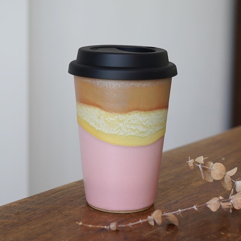 May include: A ceramic travel mug with a black lid. The mug has a pink base, a yellow and white textured middle section, and a brown top section. The mug is sitting on a wooden surface.