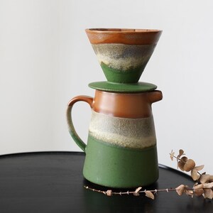 May include: A ceramic coffee pour-over set with a green, brown, and white color scheme. The set includes a carafe with a handle and a conical filter holder. The design features a layered effect with a rustic, handcrafted aesthetic.
