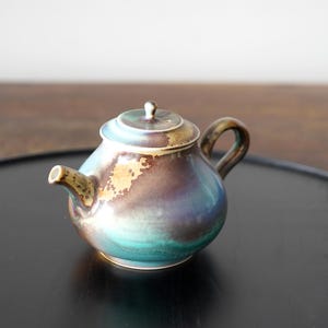 Könnte beinhalten: Eine Teekanne aus Keramik mit brauner, grüner und goldener Glasur. Die Teekanne hat einen runden Korpus und einen kleinen Ausguss. Sie steht auf einem schwarzen Tablett.