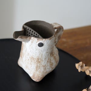 Può includere: Una brocca in ceramica fatta a mano con una superficie unica e strutturata. La brocca bianco sporco presenta un beccuccio, un manico e un interno scuro con un colino incorporato. Un piccolo punto nero è sul davanti. La brocca è progettata per versare.