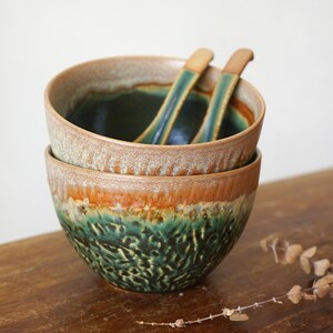 May include: Two stacked ceramic bowls with a textured, mottled glaze featuring deep forest green at the base, transitioning into a warm amber and tan rim. Two matching ceramic spoons with green and tan glaze rest inside the top bowl.
