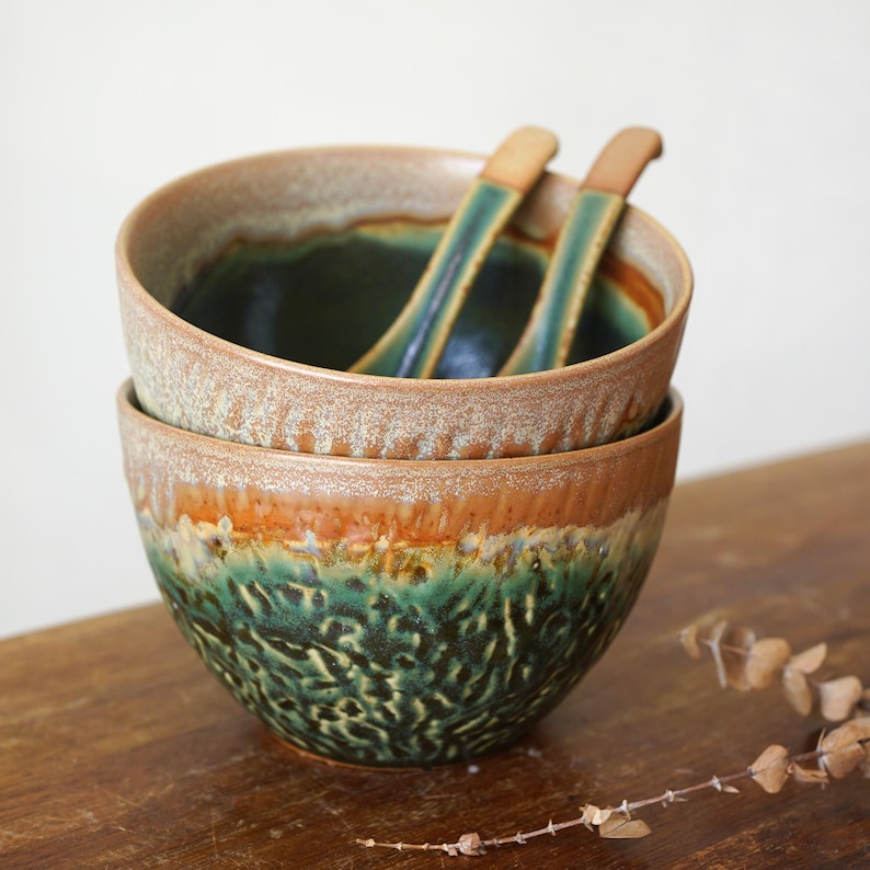 May include: Two stacked ceramic bowls with a textured, mottled glaze featuring deep forest green at the base, transitioning into a warm amber and tan rim. Two matching ceramic spoons with green and tan glaze rest inside the top bowl.