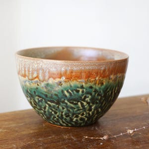 May include: A ceramic bowl featuring a reactive glaze with a smooth, light brown upper rim that transitions into a textured, mottled dark green and black base. The bowl has a rounded shape with a wide opening and a deep, curved interior basin.