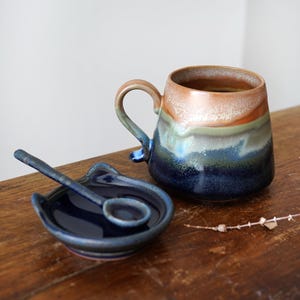 May include: A handmade ceramic mug with a matching spoon and spoon rest. The mug features a gradient of colors, including brown, green, and blue. The spoon rest and spoon are a deep blue color. The set is displayed on a wooden surface.