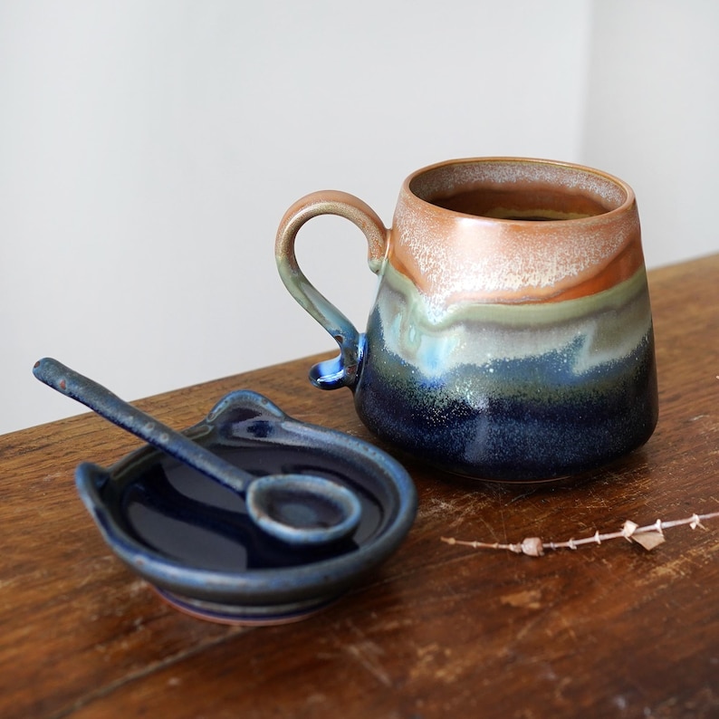 May include: A handmade ceramic mug with a matching spoon and spoon rest. The mug features a gradient of colors, including brown, green, and blue. The spoon rest and spoon are a deep blue color. The set is displayed on a wooden surface.