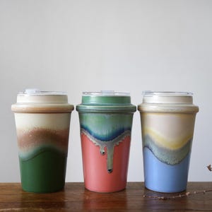 Op de afbeelding: Drie keramische reisbekers met deksels. De bekers hebben een uniek ontwerp met een kleurengradiënt, waaronder groen, bruin, rood, blauw en crème. De bekers staan op een houten oppervlak.