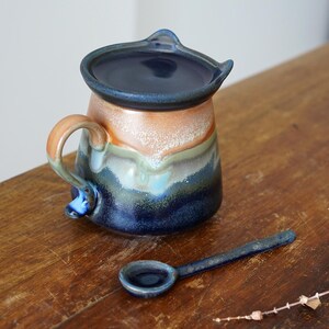 May include: A handmade ceramic creamer with a matching lid and small spoon. The creamer features a handle and spout, with a color gradient of orange, blue, and green. The spoon is dark blue.