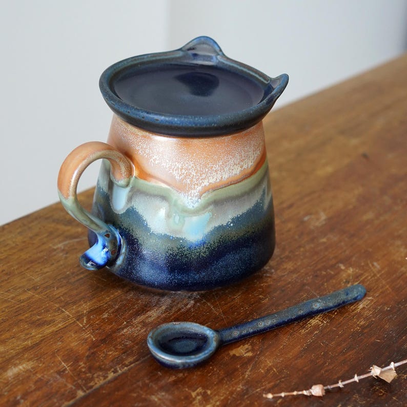 May include: A handmade ceramic creamer with a matching lid and small spoon. The creamer features a handle and spout, with a color gradient of orange, blue, and green. The spoon is dark blue.