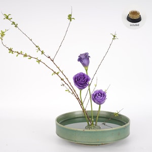 Op de afbeelding: Een groene keramische kom met water en drie paarse bloemen, gerangschikt in traditionele ikebana-stijl. De kom staat op een wit oppervlak. Een kleine, gouden bloemsteker is inbegrepen.