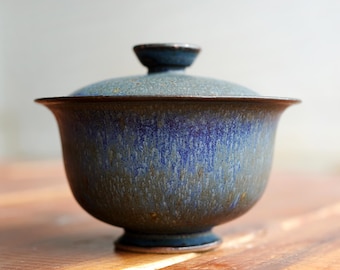 Puede incluir: Un cuenco de té de cerámica con tapa a juego, que presenta un esmalte moteado en tonos azul profundo, morado y marrón tierra. El cuenco tiene un borde ancho y acampanado, un cuerpo redondeado y una pequeña base de pedestal con acabado texturizado.