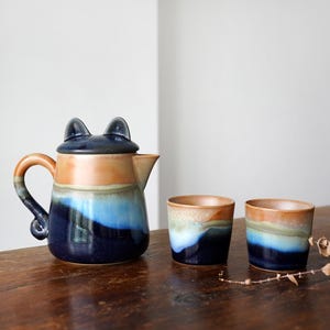 May include: A ceramic teapot with a cat-ear lid, a handle, and a spout, alongside two matching cups. The teapot and cups feature a color gradient of blue, orange, and brown. The set is on a wooden surface.